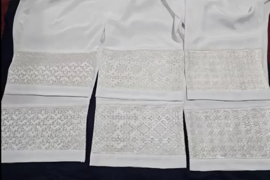 Three pairs of white trousers lie laid out on a dark surface, each with a different hand-embroidered pattern on the lower legs. (Photo credit: Social Media)