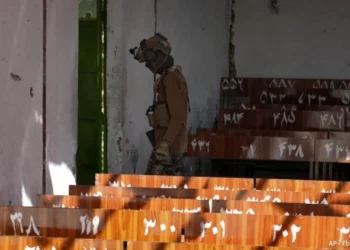 An armed soldier walks past rows of numbered wooden desks inside a damaged classroom (Photo: AP / EbrahimNoorozi)