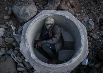 A man with his face covered by a scarf crouches inside a concrete pipe on rocky ground amid the dangers facing migrants on smuggling routes. (Photo credit: (EPA_EFE) Sedat Suna)