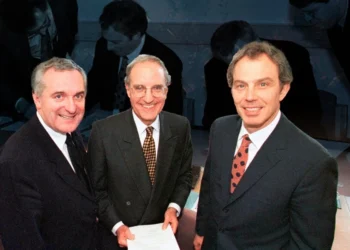 Three men in suits smile while holding a document during Northern Ireland’s peace process. (Photo credit: AP + BBC)