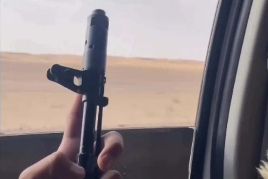 A still from a social media video shows a hand holding a rifle out of a moving vehicle’s window against a desert backdrop. (Photo credit: social media)
