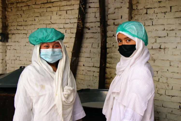 Two Afghan girls wearing face masks and protective hair coverings stand together in a food preparation area (Photo: UNDP)