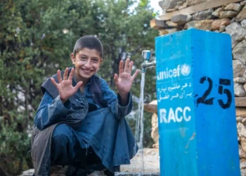 A smiling boy shows his palms while sitting beside a water tap as UNICEF supports access to safe water in Afghanistan. (Photo credit: UNICEF)